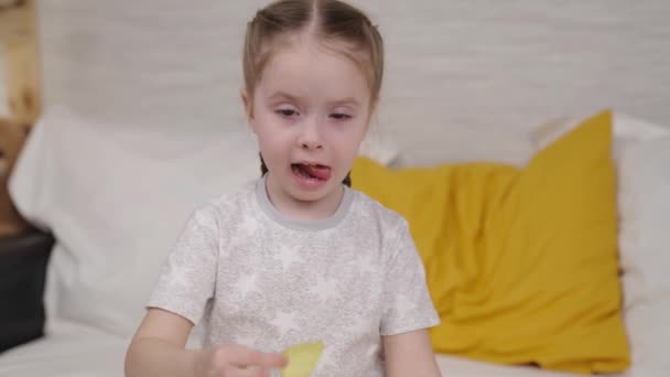 Kleines Mädchen isst goldene Kartoffelchips, die auf dem Bett im Schlafzimmer liegen, Fast Food für Kinder, ungesunde Babynahrung, genießt den Geschmack von Kartoffelgewürzen, Lieblingssnack für Mädchen zu Hause, glückliche Tochter — Stockvideo