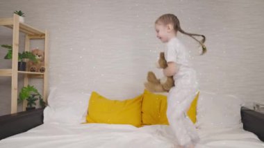 Oyuncak ayıyla yatağa atlayan neşeli küçük bir kız, evde çocuk oyunları ve eğlence, hafta sonları açık hava aktiviteleri, yatakta gülen bir çocuğun favori oyuncağı, mutlu bir aile.