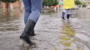 Yağmurluklu ve lastik çizmeli kızlar yağmur altında yol boyunca yürür, ayakları su birikintileri şehrinde yürür, kenarlara su sıçratır, sel caddede, araba su üstünde gider.