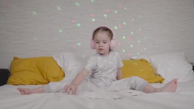 Kulaklıklı küçük çocuk müzik dinler ve yatağın üzerinde bozuk paraları sayar, duvarda çok renkli bir çelenk parıldar, çocuklar için para hayal eder, kız çocuğunun müziğini dinler.