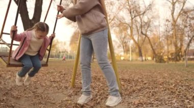 Küçük bir çocuk salıncakta sallanıyor ve uçarken gülüyor, bir sonbahar oyun parkı, mutlu bir aile, bir anne çocuğunu şehir parkında yuvarlıyor, çocukluk hayali uçuyor, annesine yürüyüş yaparken gülümsüyor, bir bebekle oynuyor.