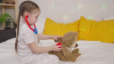 Küçük çocuk hastanede oyuncak ayıyla oynuyor, çocuk steteskoplu oyuncak dinliyor, çocukluk hayali doktor oluyor, bebek kendini veteriner olarak hayal ediyor, kız hastayı muayene ediyor.