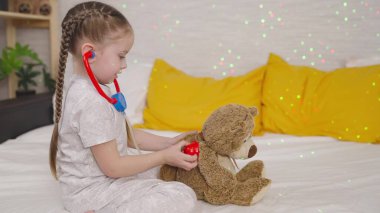 Küçük çocuk hastanede oyuncak ayıyla oynuyor, çocuk steteskoplu oyuncak dinliyor, çocukluk hayali doktor oluyor, bebek kendini veteriner olarak hayal ediyor, kız hastayı muayene ediyor.