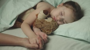 Küçük çocuk yatakta uyuyor. Oyuncak ayıya sarılıyor. Annesinin elini tutuyor. Mutlu bir aile..