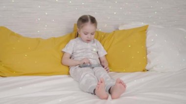 Küçük kız internet üzerinden akıllı telefon uygulaması kullanarak öğreniyor, okul platformunda uzaktan öğreniyor, atamalar yapıyor, çocuklar oyun oynuyor modern telefon boyalı ışık, çocuk yatağında oturuyor.