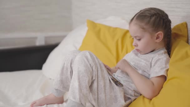 Small child is playing game on modern smartphone while sitting on bed in a bedroom, kid is studying in the phone application, doing baby homework online, watching TV entertainment programs on gadget — Stock Video