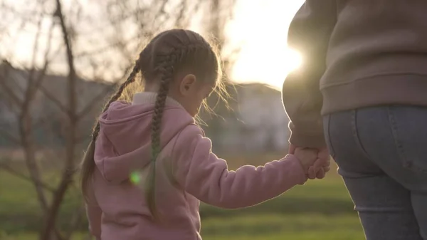 Criança segura a mão de suas mães nos raios de luz no parque, menina vai passear com sua mãe para o playground, conceito de vida familiar feliz, criança ao pôr-do-sol da noite com um adulto é alegre — Fotografia de Stock