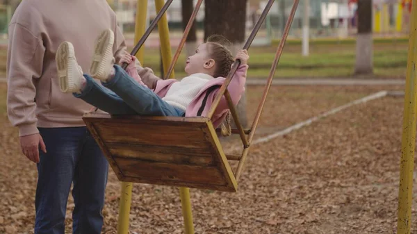 Mother shakes little child on playground swing, cheerful kid flies up and down, baby laughs and smiles while playing, mother and daughter on walk in city park, happy family, childhood dream of flying — Stock Photo, Image