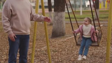 Anne küçük çocuğu sallıyor salıncakta, neşeli çocuk yukarı aşağı uçuyor, bebek oyun oynarken gülüyor ve gülümsüyor, anne kız parkta yürüyor, mutlu aile, çocukluk hayali uçuyor.