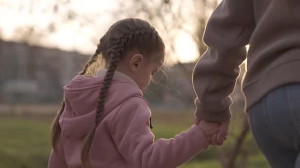 Child holds her mothers hand in the rays of light in the park, little girl goes for walk with her mother to playground, concept of happy family life, kid at evening sunset with an adult is cheerful — Stockvideo
