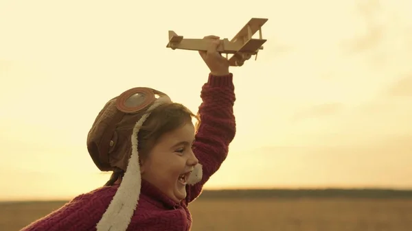 Little happy girl runs with a wooden plane in hand at sunset in sky and laughs, running child plays the game flying over ground, kids runner outdoors, kid flyer dream on vacation travel, baby weekend — Zdjęcie stockowe
