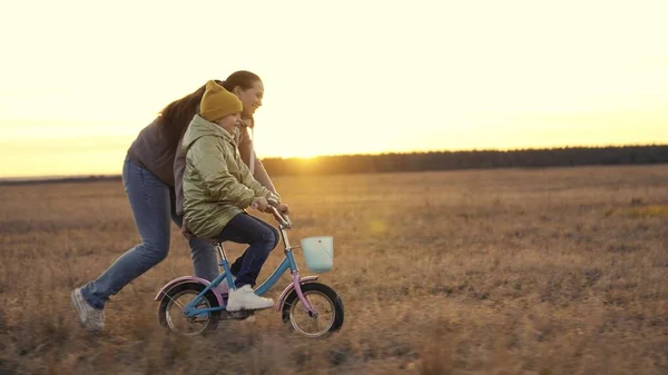 Mother teaches little daughter ride two-wheeled bike at sunset, child pedals on bicycle in sunshine, outdoor outdoor activities, exercise at dawn travel, happy family life, childhood dream drive — Stockfoto