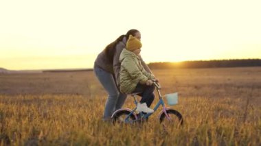Anne küçük kızına gün batımında iki tekerlekli bisiklete binmeyi öğretiyor, çocuk pedalları güneş ışığında bisiklet sürüyor, açık havada aktiviteler yapıyor, sabah gezisinde egzersiz yapıyor, mutlu aile hayatı, çocukluk rüya sürüşü.