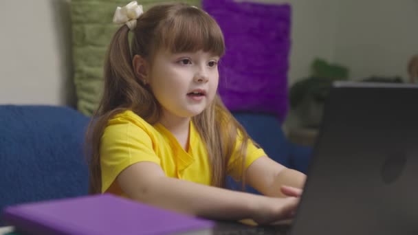 Niño pequeño hace la tarea en un ordenador portátil, una chica escribe en un teclado, la escuela en casa en un ordenador en línea, el aprendizaje de un niño de forma remota en casa, jugando juegos en el programa de aplicaciones informáticas, la tecnología moderna — Vídeo de stock
