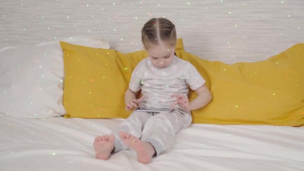 Small child plays in smartphone display, a child does lessons in a smartphone application remotely at home, lying on a bed, a girl reads online gadget chat correspondence, children books on Internet — Stock Video