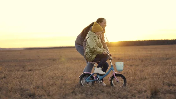 Mãe ensina pequena filha andar de bicicleta de duas rodas ao pôr do sol, pedais de criança na bicicleta ao sol, atividades ao ar livre, exercício ao amanhecer viagens, vida familiar feliz, unidade de sonho da infância — Fotografia de Stock