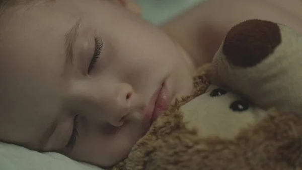 Малыш спит, обнимая плюшевого медведя, лежа в постели, мальчику снится мягкая игрушка, накрытая теплым одеялом в детской комнате, закрытые глаза детского отдыха по ночам, дремлет в темноте с игрушечным другом — стоковое фото