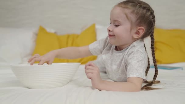 Girl eats potato chips lying bed and laughs, quick meal for kid, small child snacking on crispy potatoes, good appetite of cheerful child, fatty high-calorie fast food of carbohydrates, enjoy taste — Stock Video