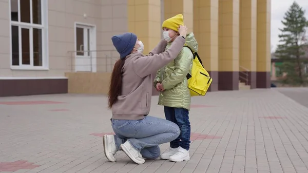 Matka si nasazuje na obličej lékařskou masku pro školačku, aby chránila malé dítě během školních hodin, matka objímá dítě s batohem, ochrana před virovou chorobou koronavirové infekce, covid-2019 — Stock fotografie