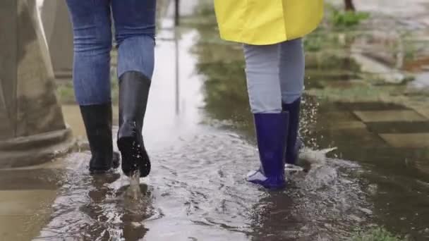 Niñas en impermeables y botas de goma caminan a lo largo del camino inundado de lluvias torrenciales, sus pies caminan a través de la ciudad charcos, salpicaduras de agua a los lados, la inundación está en la calle, el coche está conduciendo en el agua — Vídeos de Stock