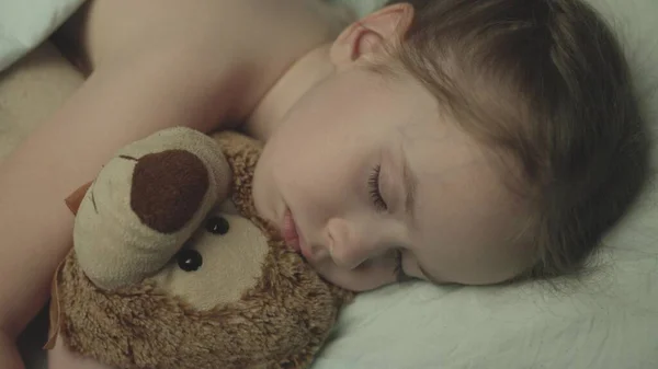 Malé dítě spí v noci v postýlce objetí s plyšovým medvídkem, dívka dcera sny při ležení postel se zavřenýma očima, dítě s kamarádkou s panenkou medvídka odpočinku spolu, dítě tvář a hračky zblízka — Stock fotografie