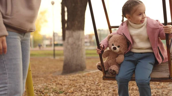 Un niño pequeño se balancea con un oso de peluche en un parque de otoño, un niño feliz sueña con volar con su juguete favorito, unas vacaciones familiares activas de madre e hija a pie en la ciudad, amigo de la infancia — Foto de Stock