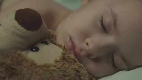 Kid sleeps lying in bed hugging a teddy bear, close-up of babys face with closed eyes, child night sleep with dreams, tired child napping in bedroom, time for night rest with friend doll together — Fotografia de Stock