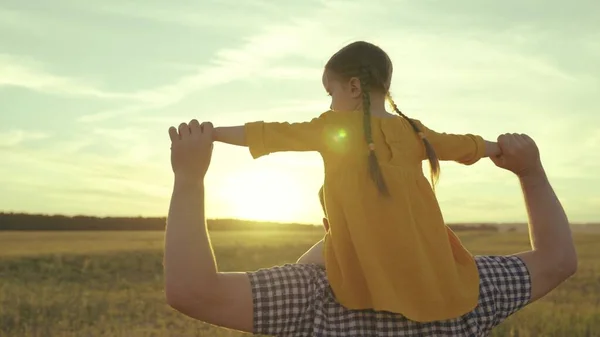 Małe dziecko siedzi na ramionach ojców z podniesionymi dłońmi o zachodzie słońca, córeczka z tatą na spacerze, szczęśliwe życie rodzinne, marzenie z dzieciństwa, aby latać, być pilotem, dziewczyna z tatusiem w promieniach słońca, bawić się — Zdjęcie stockowe