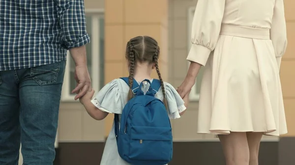 Liten flicka går hand i hand med föräldrar till skolan, unge med en ryggsäck på ryggen, undervisning i grundskolan, undervisning i skolans utbildningsprogram, lycklig familj — Stockfoto