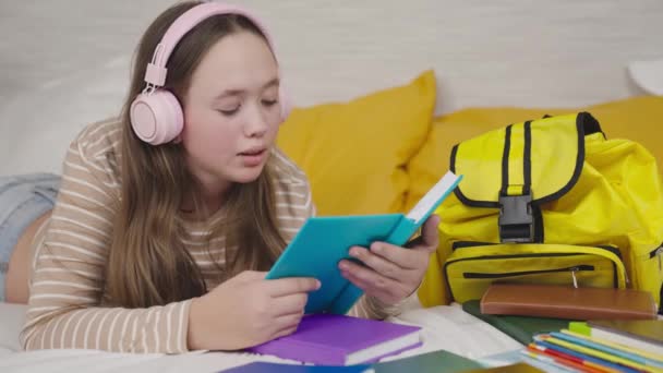 Dospívající dívka leží na gauči a čte si knihu, dítě poslouchá hudbu na sluchátkách, moderní dítě studuje školní literaturu, vzdělávání mladých studentů, získává znalosti prostřednictvím stránek s knihami — Stock video