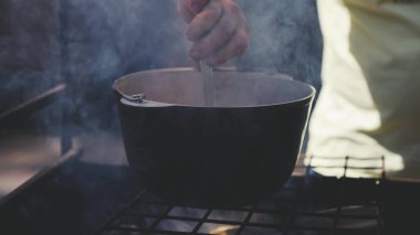 Bir adam ateşte yemek pişiriyor, turist kampında tencerede lezzetli yemekler pişiriyor, dumanlı kömürlerin tarifi, ormana yolculuk, aktif olarak geçirilen bir hafta sonu, kaşıkla yemek pişiriyor.