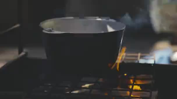Un hombre cocina comida en una olla en llamas, deliciosa cocina en una cacerola en un campamento turístico, una receta sobre carbones en humo, un viaje al bosque a la naturaleza, un fin de semana activamente pasado, cocinar revuelve la comida con cuchara — Vídeo de stock