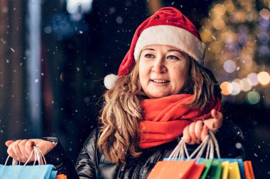 Noel Baba şapkalı bir kadının portresi Noel ışıkları ve süslemelerle süslenmiş renkli çantalarla Noel alışverişi yapıyor..