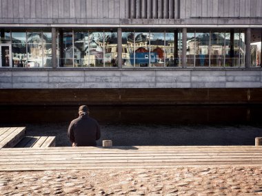 Bir adam rıhtımda tek başına oturuyor. Şehrin renkleri karşı binadaki pencerelere yansıyor. Bir sanat galerisine benziyor..