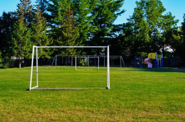 Okul bahçesinde boş bir futbol sahası, sabah güneşinde beyaz hedefler ve arka planda ağaçlar