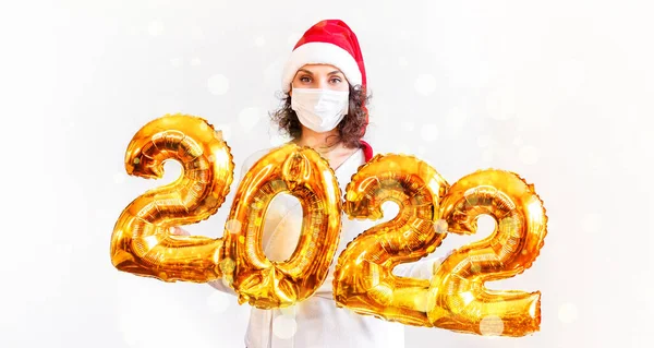 Jeune belle fille en chapeau de Noël rouge et masque médical tenant des ballons 2022 dans ses mains. Concept de célébrer le nouvel an au moment de la menace covid-19. Nouvel An 2022 or Nombre ballons — Photo