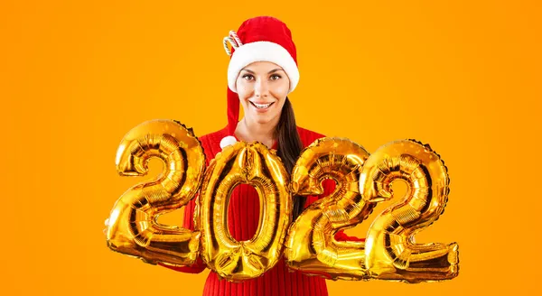 Nouvel An 2022 ballons d'or. Jeune femme en chapeau de Père Noël ayant souriant tenant dans la main 2022. Fille en chapeau rouge et robe rouge se tient sur fond orange vif. Concept Fête de Noël — Photo