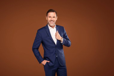 Confident elegant handsome business man standing in front of a brown studio background, wearing fashionable suit, smiling and looking at the camera clipart