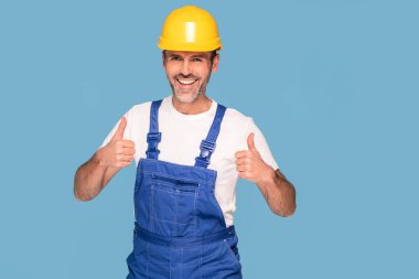 Sarı kask takan yakışıklı bir endüstri mühendisi, mavi pastel stüdyo arka planında izole edilmiş. Mutlu işçi gülümsüyor ve kameraya bakıyor.