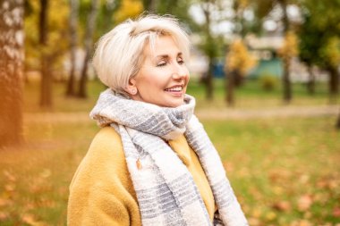 Parkta rahatlatıcı bir yürüyüşe çıkmış neşeli yetişkin bir kadın. Gülümseyen güzel bayan sonbahar mevsiminde güneşli havanın tadını çıkarıyor.. 