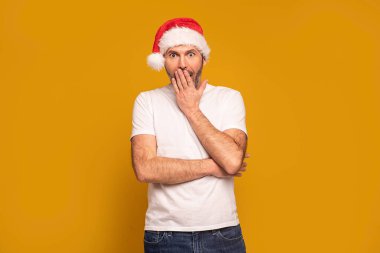 Etkilenmiş bir adam ellerini çenesine koyuyor, doğrudan kameraya bakıyor, kırmızı Noel Baba şapkası takıyor, şok oluyor, renkli stüdyo arka planında izole edilmiş poz veriyor. Xmas zamanı..