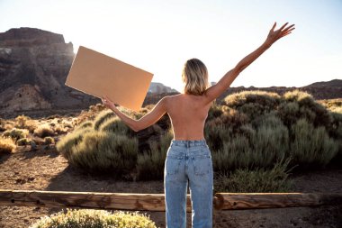 Kot pantolonlu kadın, üstsüz, özgürlük ve eşitlik için protesto eden, elinde fotokopi alanı olan boş pankart tutan. Elleri havada bir kız. Arkadan bak. Vahşi doğa. Kadın iktidarı, protesto, kadın hakları, aktivizm konsepti.