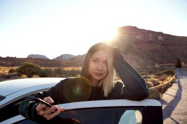 Çevrimiçi haritalı akıllı telefon kullanan mutlu bir kadın gün doğumunda ve off-road gezisinde dağlık arazide araba yolculuğunun tadını çıkarıyor. Gerçek insan yaşam tarzı.