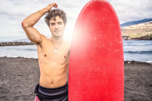 Young handsome spanish man with curly hair standing shirtless on the beach with red surfboard, smiling to the camera. Good vibes only. Summer time. Hobby. Water sport