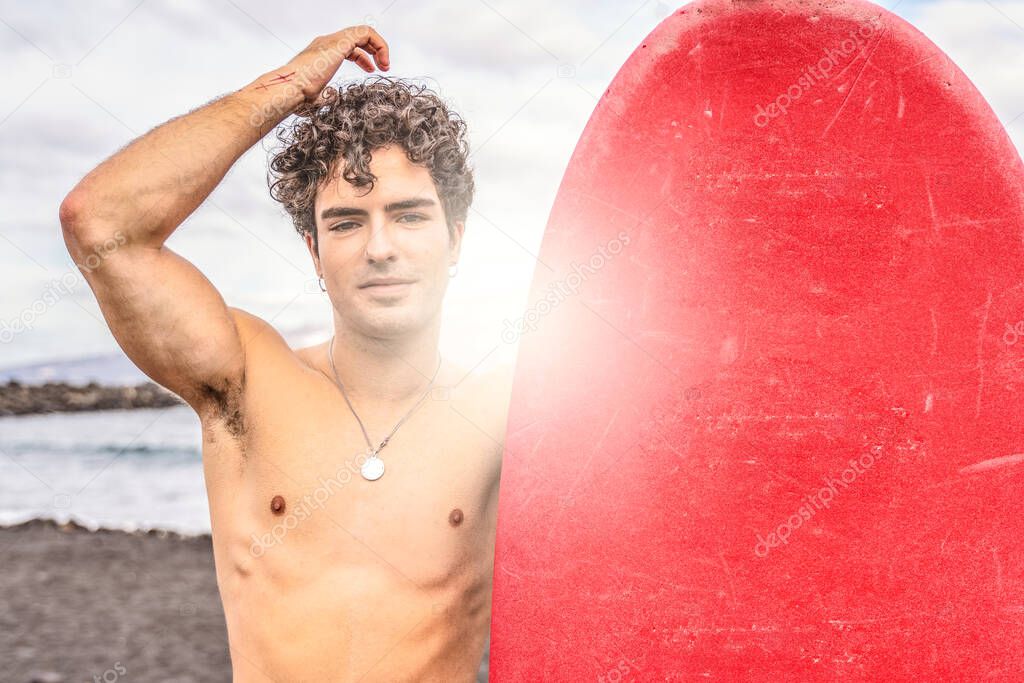 Young handsome spanish man with curly hair standing shirtless on the beach with red surfboard, smiling to the camera. Good vibes only. Summer time. Hobby. Water sport