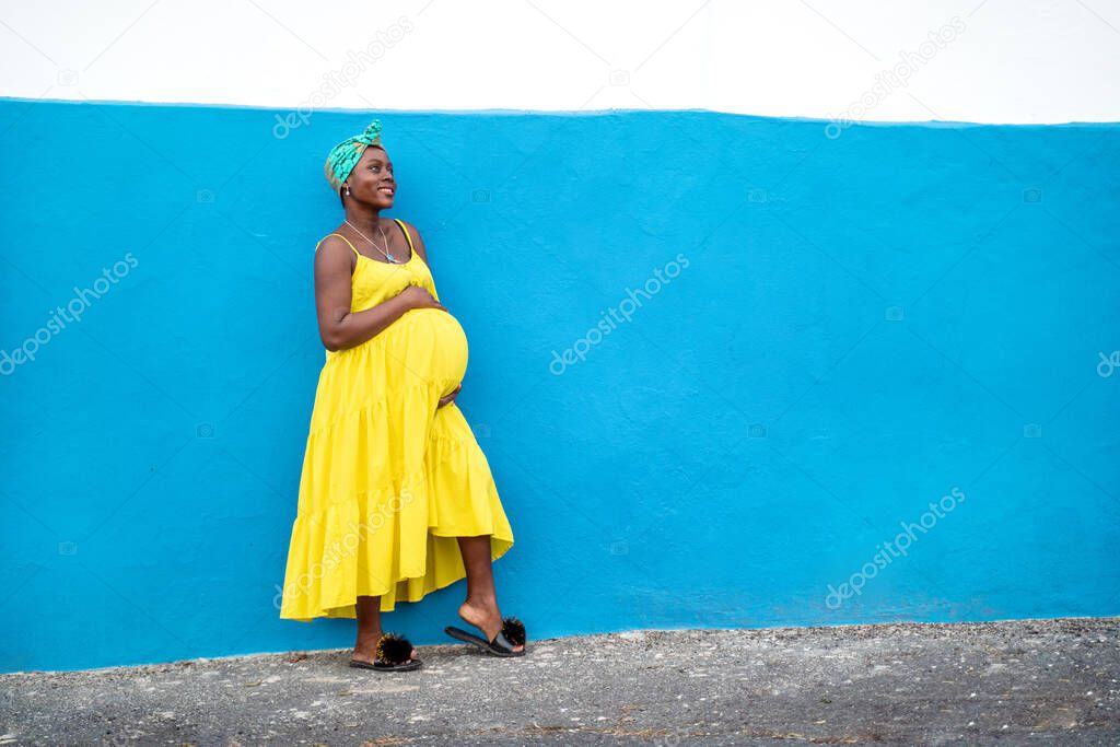 Happy pregnant african woman in yellow dress holding her belly in hands, smiling, posing on the blue wall outdoor. Real people lifestyle and emotions. Maternity. Pregnancy