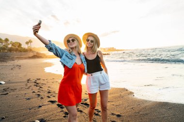 Güzel günbatımında güzel bir telefonla sahilde selfie çeken mutlu sarışın beyaz bayan arkadaşlar. Rahat ol. Özgürlük için. Mutluluk. Arkadaşlık. Kız kardeşler. Gerçek insanların duyguları..