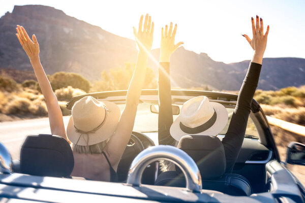 Dreams come true! Two happy young girls driving cabrio car during vacation road trip in mountains, making memories and having fun together. Back view. Hands up. Freedom concept. Happiness. Tourist. Wanderlust. Real people emotions.