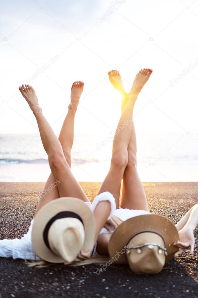 Two female friends, sisters lying on the bach in summer hats and with legs upside down, relaxing, sunbathing. Sunset Time. Vacation concept by the sea. Friendship. Lifestyle. Tourism. Tourist