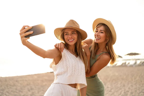 两个快乐的金发美女夏天在海滩上玩得很开心 带着手机自拍 笑着看着智能手机摄像头 戴帽子的漂亮姑娘 真实的人的情感 — 图库照片